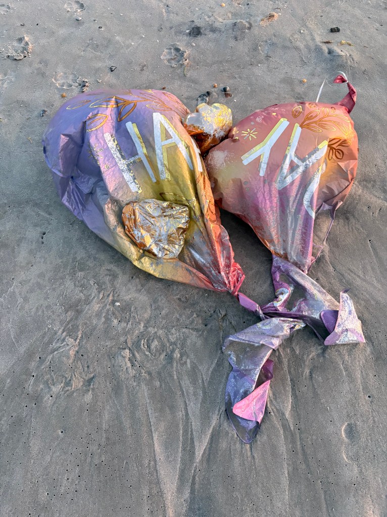 Two pastel gradient mylar balloons tangled on the beach, partially buried in sand