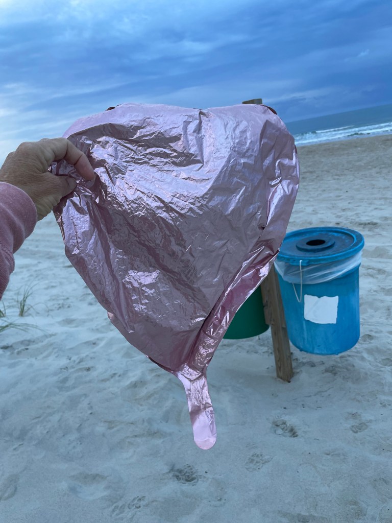 A light pink mylar balloon being held up in front of the beach on a cloudy day.