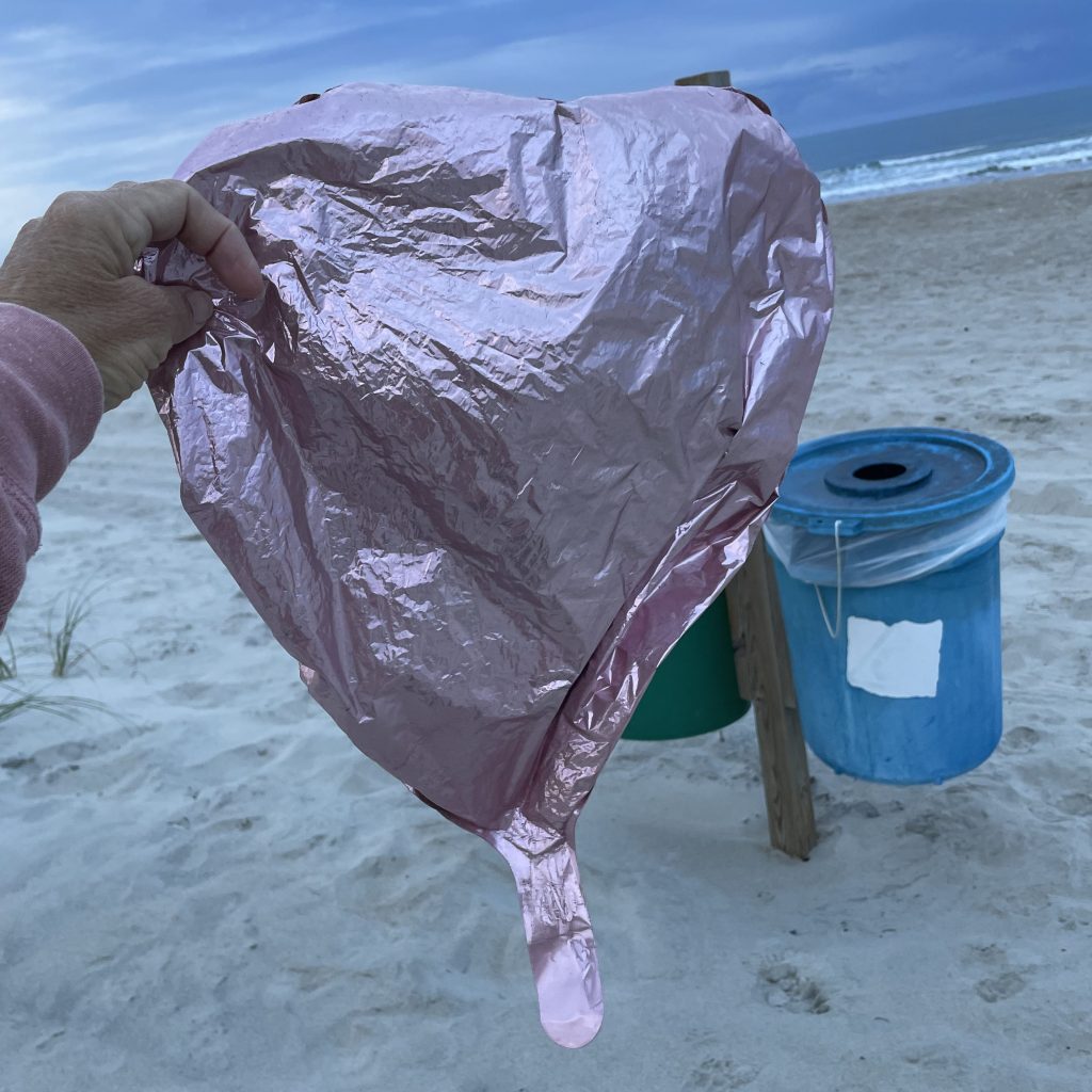 A light pink mylar balloon being held up in front of the beach on a cloudy day.